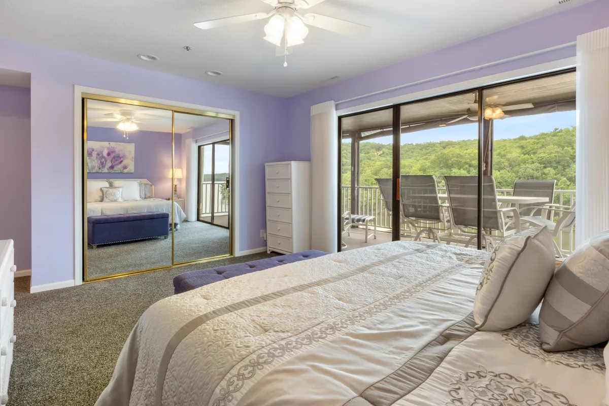 A serene bedroom with a large bed, decorative pillows, sliding glass door to a balcony, and a mirrored wardrobe, offering an inviting and safe retreat within the condo.