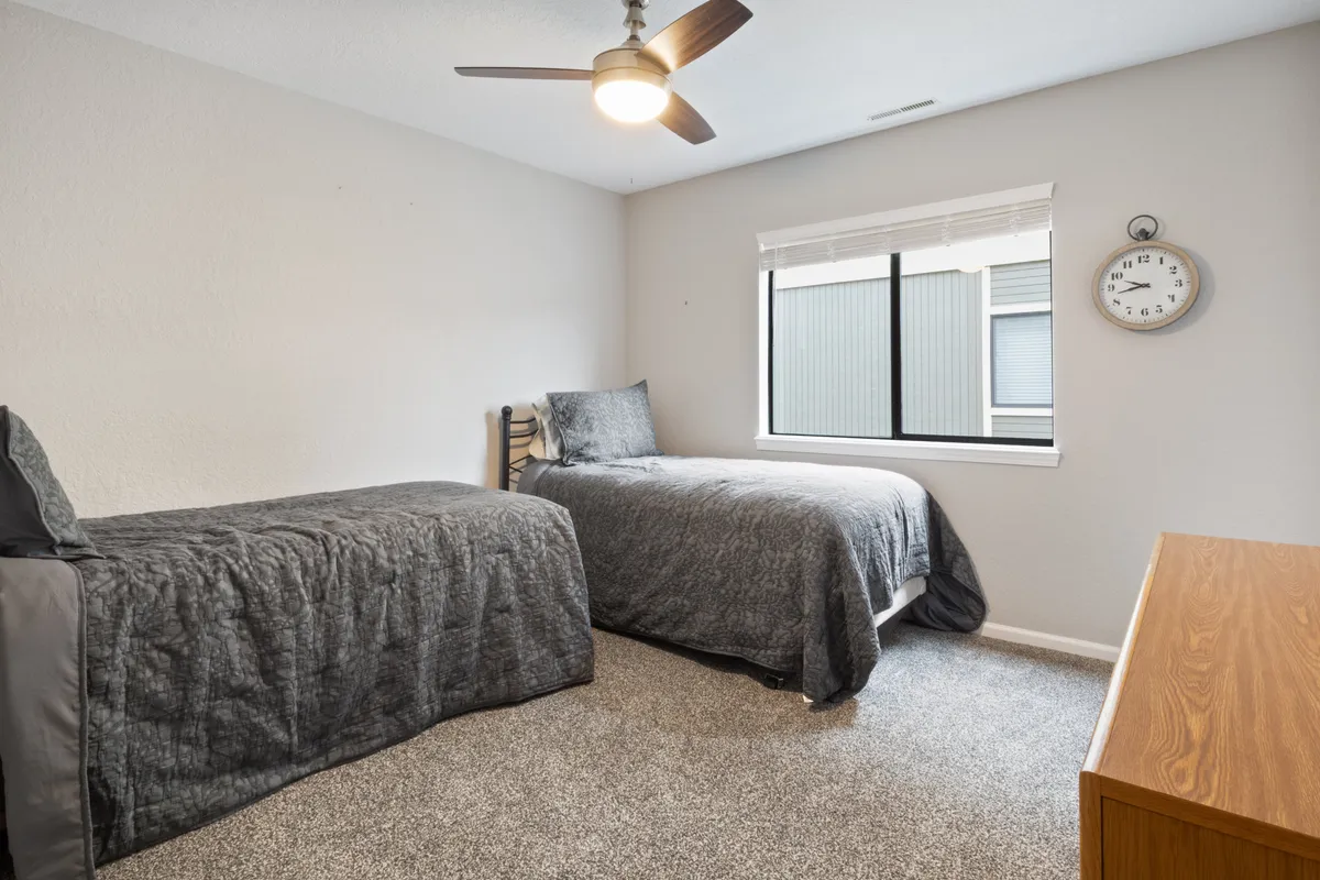 A serene bedroom featuring two twin beds with crisp white bedding, a large window letting in natural light, a wooden dresser, and a classic wall clock. The space feels airy and welcoming, perfect for children or guests traveling together.