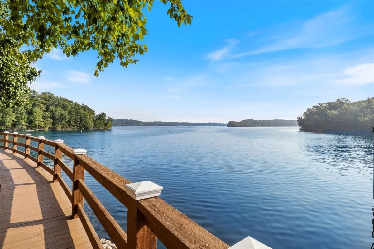 A tranquil lakeside view with a wooden railing in the foreground, surrounded by lush trees and an open sky. The scene is peaceful and inviting, suggesting pathways perfect for morning walks, fishing, or simply enjoying the natural setting with your pet.