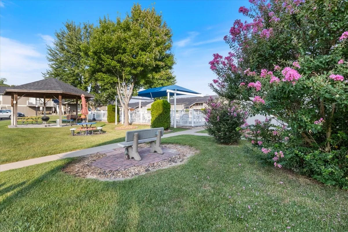 A peaceful outdoor setting with benches, flowering shrubs, a gazebo, and picnic tables. The area feels calm and natural, offering shaded spots for relaxation, socializing, or letting a pet enjoy some fresh air.