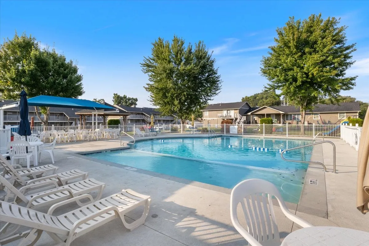 A cheerful pool area surrounded by lush trees, featuring a sparkling swimming pool, numerous lounge chairs, and a modern shade structure. The environment is ideal for both lively summer pool parties and peaceful afternoons spent sunbathing or relaxing.