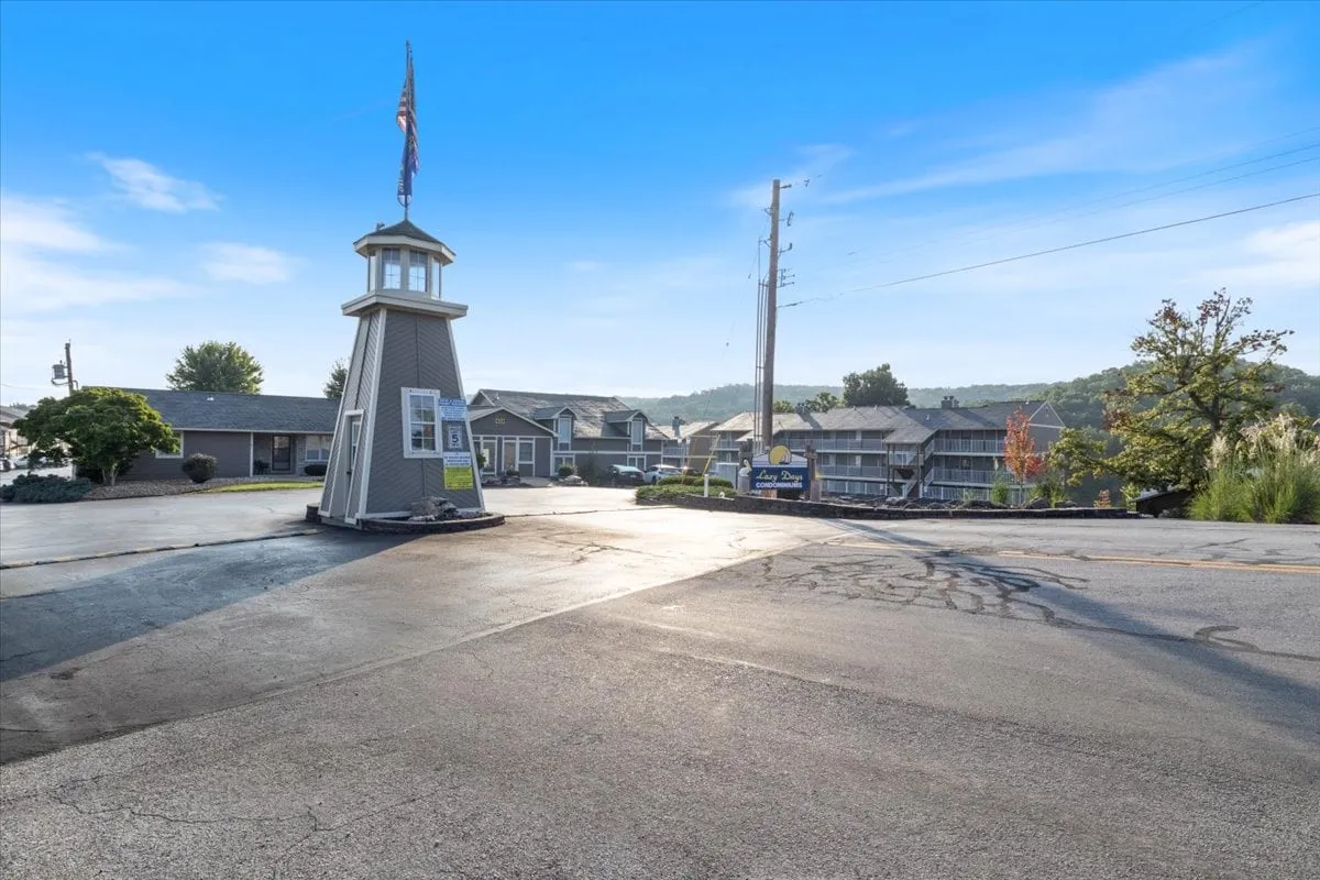 The welcoming entrance to the property is marked by a distinctive lighthouse structure, a flag pole, and a landscaped roadway leading into the area. The surrounding landscape changes with the seasons, hinting at the vibrant community atmosphere.