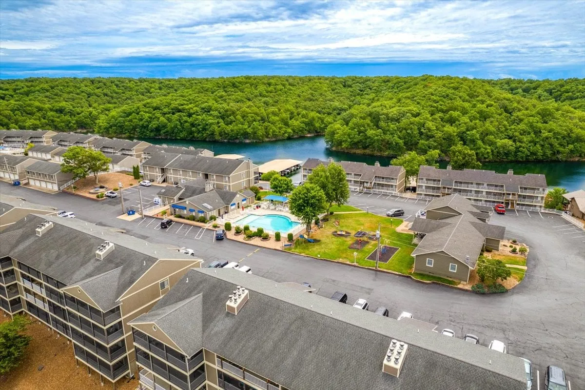 A bright and welcoming exterior scene featuring a sparkling swimming pool, lush greenery, and the nearby river, with a parking area visible in the background. The pool area is designed for relaxation, swimming, picnicking, and enjoying the surrounding natural beauty.