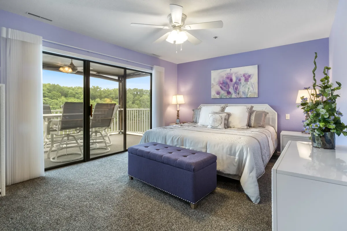 A king-sized bed with plush bedding is set against a purple accent wall, complemented by glass sliding doors that lead to a private balcony. A decorative bench sits at the foot of the bed, while nightstands and plants add warmth and coziness to the space. Natural light streams in, highlighting the tranquil and comfortable ambiance.
