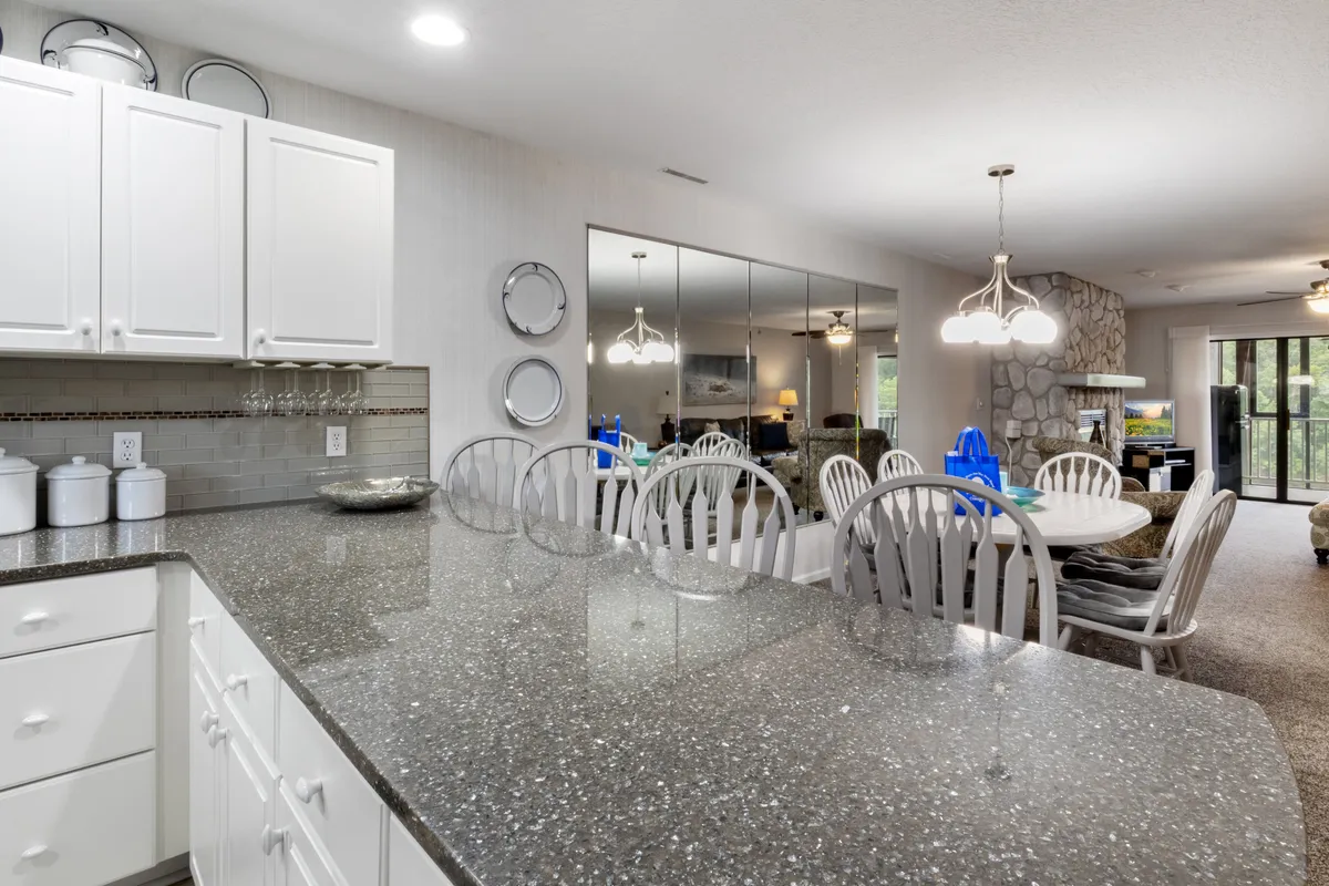 This image captures a bright, open-concept space that seamlessly blends the kitchen, dining table, and living area. The dining table is set near a kitchen island, with a glimpse of a stone fireplace anchoring the cozy living space visible in the reflection. The overall atmosphere is lively and communal, perfect for gatherings and shared meals.